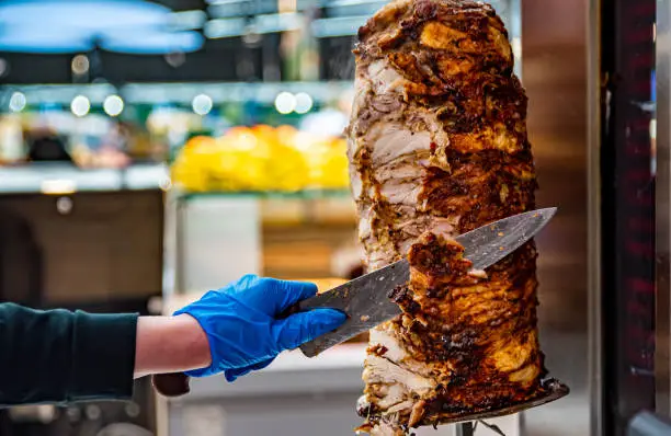 hands holding a knife, cutting slices from a vertical skewer. Doner Kebab