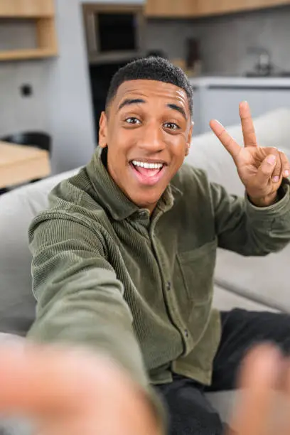 Photo of Great selfie. Multiracial young guy gesturing to the smartphone camera, while making funny photos at home