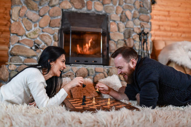 Couple Playing Chess And Warming Up By Fireplace In Mountain Cottage
