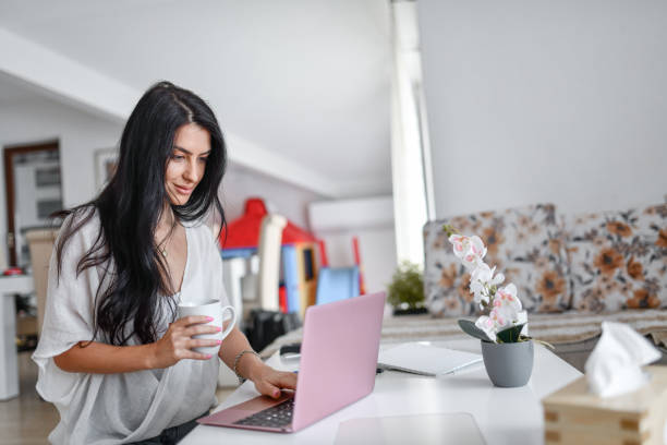 istock Beautiful Female Drinking Cup Of Coffee While Using Laptop Home 1368432383