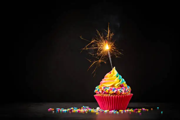 Rainbow Birthday cupcake with a sparkler and colourful sprinkles over a dark background. A magical Birthday celebration.