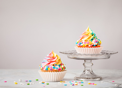 Two Rainbow Birthday cupcakes with colourful sprinkles over light grey background.