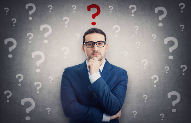 istock Skeptical businessman surrounded by interrogation marks has a main question as a red symbol over head. Thoughtful business person keeps hand under chin, waiting for an answer 1368440123