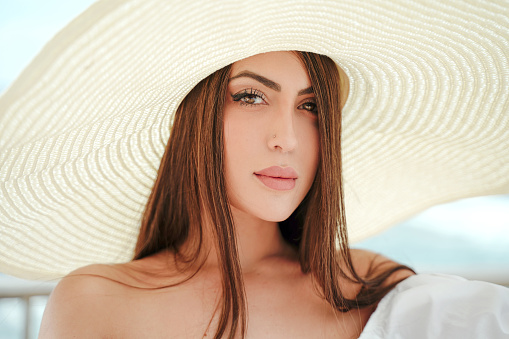 Closeup beauty portrait of young attractive brunette woman with long hair and brown eyes, wearing big summer hat, looking at the camera. Outdoor sunny photo.