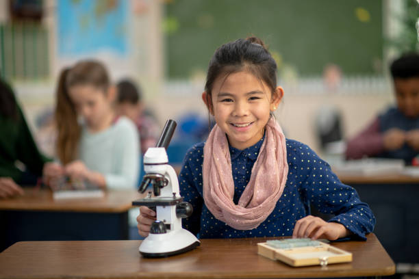 A sweet little girl of Filipino decent, is seen sitting at her desk with a microscope out in front of her as she works away in science class.  She is dressed casually and looking up with a smile as she adjusts the microscope before analyzing the slide on it.  Her peers can be seen working away in the background at their individual desks as well.