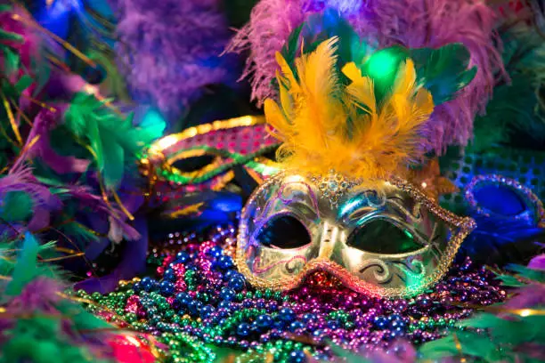 Photo of LV Mardi Gras sequined mask, decorated with feathers on a bed of feathered Mardi Gras feather Boas.  Colorful beads and spot lights of color.
