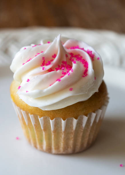 A close up view of a mini cupcake decorated with pink sugar sprinkles.  Confetti cupcake batter with white frosting on a white plate.  Wooden table blurred in background.