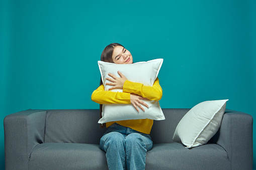 Happy young girl hugging pillow sitting on sofa at cozy home, resting, relaxing on blue background. Biorhythms, healthy sleep concept. High quality photo