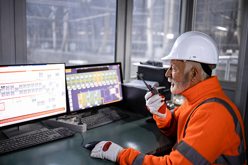 Industrial worker in safety equipment sitting in factory control room monitoring production process and talking on radio communication.
