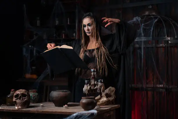 Halloween concept. Witch dressed dark dungeon room use magic book for conjuring magic spell. Female necromancer wizard gothic interior with skull, cage, spider web