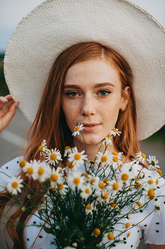 Close-up portrait of a redhead woman in hat and holding a bouquet of daises in sunny summer day. Woman in camomile field. Natural beauty. Happy people.