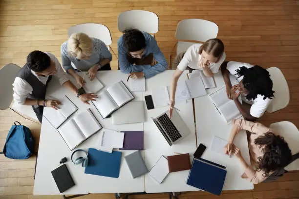 Team of young college students working on study project in university library, using laptop, writing notes at table with books, sharing learning tasks. Top aerial view