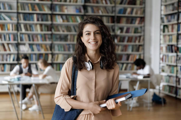 Happy Hispanic gen Z student girl with headphones visiting library Happy Hispanic gen Z student girl with headphones visiting public library for work on study research project, holding learning papers, notebook, looking at camera, smiling. Head shot portrait 20 29 years stock pictures, royalty-free photos & images