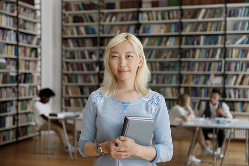 Confident positive Asian blonde college girl standing in university library with bookshelves behind, holding books, looking at camera, smiling, Fresh female student head shot portrait