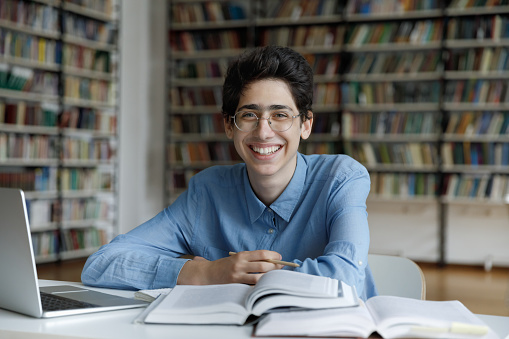Happy joyful fresh student guy in glasses writing essay in library, working on research study project, using pile of books, laptop, writing notes, looking at camera, smiling. Head shot portrait