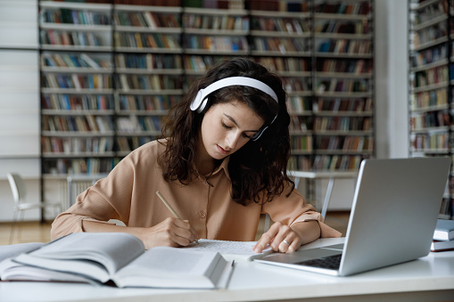 Serious busy hardworking student girl in headphones working essay, study project in college public library, attending online learning conference, watching video lesson on internet, writing notes