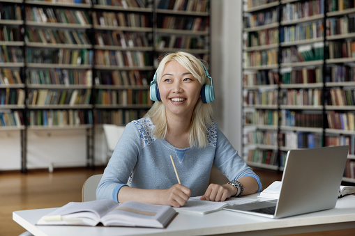 Happy pensive Asian college student girl doing homework in public library, using laptop, wireless headphones, writing notes, looking away, thinking over ideas for research article, essay