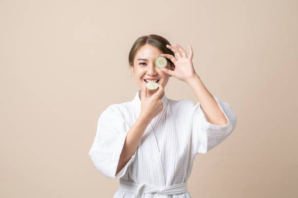 Portrait Beautiful young Asian women in bathrobe with holding cucumber slice for facial face mask on brown isolated background. stock photo
