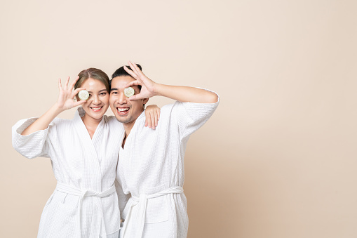 Happy Asian couple smile in bathrobe or spa suit with cucumber slice for facial mask together on brown isolated studio background.