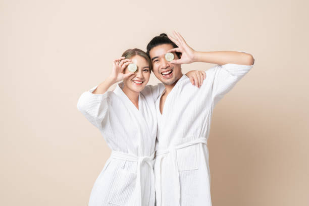 feliz pareja asiática en bata de baño con máscara facial de rodaja de pepino juntos sobre fondo marrón aislado. - facial mask spa treatment cucumber human face fotografías e imágenes de stock