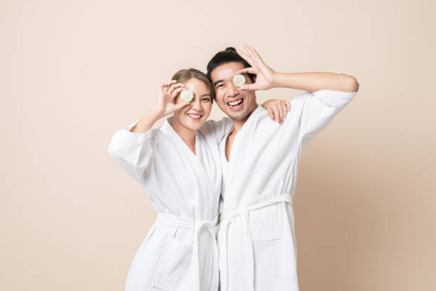 Happy Asian couple in bathrobe with cucumber slice facial mask together on brown isolated background. Happy Asian couple smile in bathrobe or spa suit with cucumber slice for facial mask together on brown isolated studio background. japanese girlfriends stock pictures, royalty-free photos & images