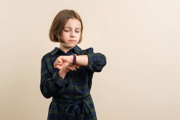 Disgruntled Kid Little Girl 9s Wearing Plaid Shirt Dress looks at a smart watch fitness band. Children time management concept