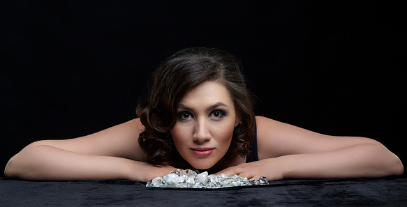 Portrait of beautiful long-haired brunette woman on black background. Female sit at table covered by black velvet. There are glass stones on the table.