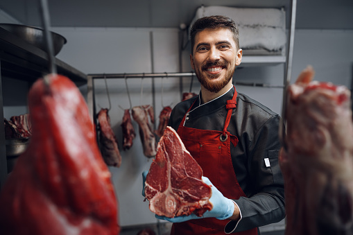 Young butcher holding raw meat steaks in fridge of grocery shop, close up