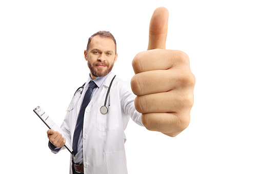 Young male doctor holding a clipboard and gesturing thumbs up in front of camera isolated on white background