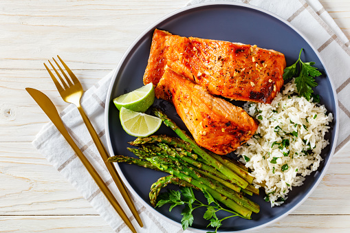 fried salmon fish fillets with asparagus, jasmine rice and lime on a plate, on a white wooden table with cutlery, horizontal view from above, flat lay, close-up