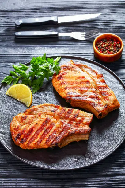 grilled bone-in pork chops on a black plate with lemon slice and parsley on a wood table with cutlery, vertical view from above
