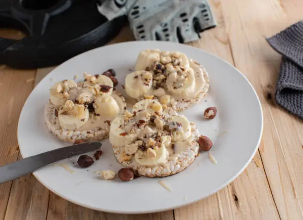 Delicious fitness breakfast for energy or muscle building with brown rice cracker topped with almond butter, fresh bananas and hazelnuts. Served on a white plate on wooden table with weight plate
