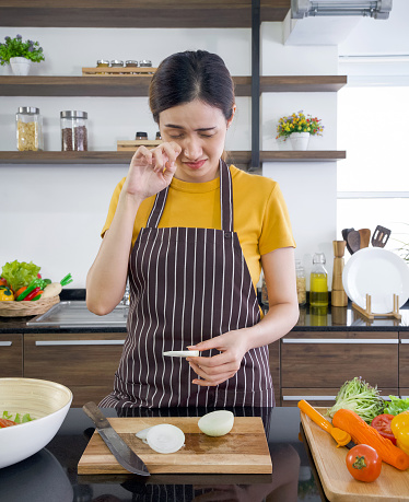 Asian woman crying while holding a piece of onion. Young housewife slices onion into pieces on a wooden chopping board. Morning atmosphere in a modern kitchen.