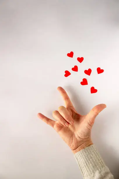 The phrase I-Love-You in sign language, the language of the deaf. Woman's hand on a white background with red paper hearts, copy space.