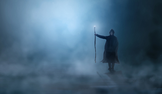 Mago en capa, capucha con palo mágico de pie en un paisaje de niebla iluminado por la luna azul ligh. Fantasía, concepto de mago, renderizado 3D photo