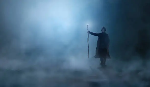 Photo of Magician in cloak, cowl with magic stick standing in fog landscape illuminated by blue moon ligh. Fantasy, wizard concept, 3D rendering