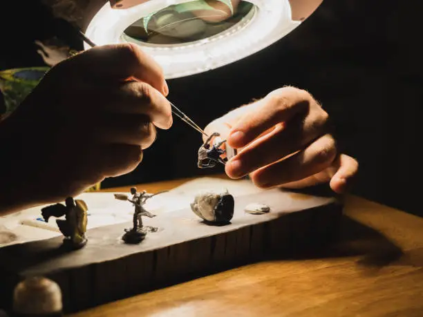 Photo of human hands painting under the lamp with light and magnifying glass plastic minifigures for board games , modern hobby of game collector
