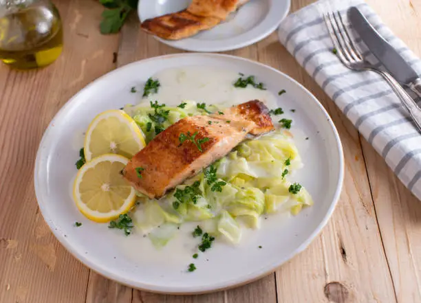 Fresh and homemade cooked fish dish with pan fried salmon fillet served with white cabbage and bechamel sauce on a plate on wooden table background . Ready to eat