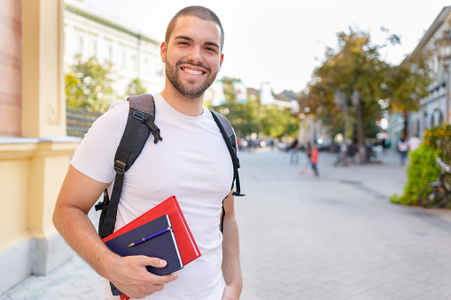 Low angle of positive young bearded male student in casual clothes with backpack and notebook smiling and looking at camera while standing on stairs of university building