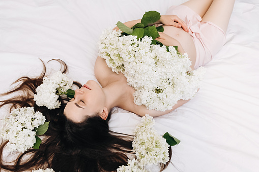 Charming nice pretty pregnant woman lying on cozy bed with beautiful spring flowers. Portrait of dreamy young brunette female 20s with eyes closed. Hair Care, skin beauty health care concept
