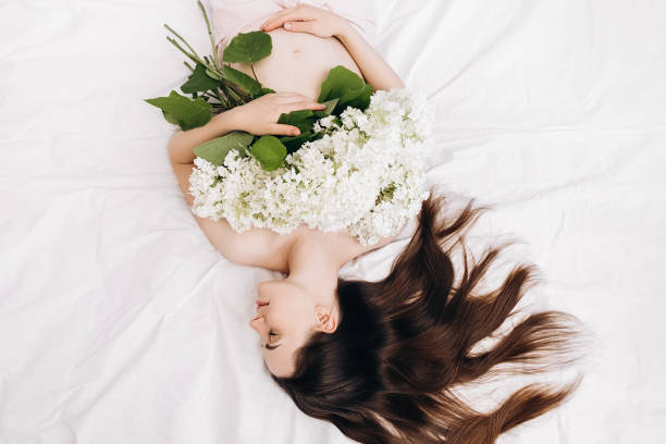 Charming attractive pregnant young female gently holds hands on tummy lying on comfy white bed at home with spring bouquet flowers. Concept motherhood and maternity. Future mother's expectation encantadora y atractiva hembra joven embarazada sostiene suavemente las manos sobre la barriga acostada en una cómoda cama blanca en casa con flores de ramo de primavera. concepto maternidad y maternidad. la expectativa de la futura madre