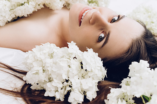 Side view beauty portrait of attractive beautiful woman with well-groomed skin and sincere smile lying on cozy white bed near white flowers around head. Cute brunette girl happy looking at camera.