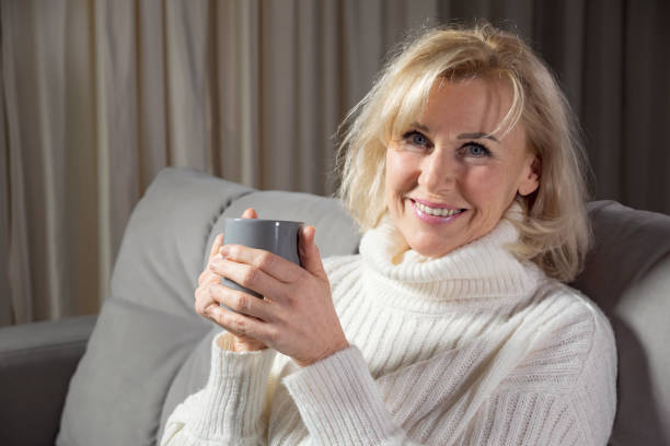 Mature woman with blonde hairstyle and mug of coffee smiles and looks at camera sitting on sofa in living room, Mature woman with blonde hairstyle and mug of coffee smiles and looks at camera sitting on sofa in living room, closeup curtain bangs stock pictures, royalty-free photos & images