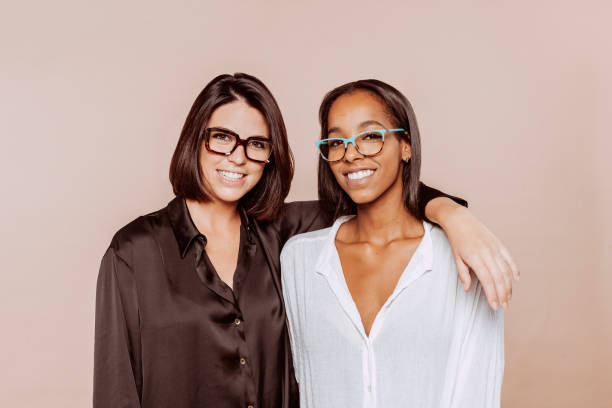 Two women with eyeglasses embrace and smile positively at studio