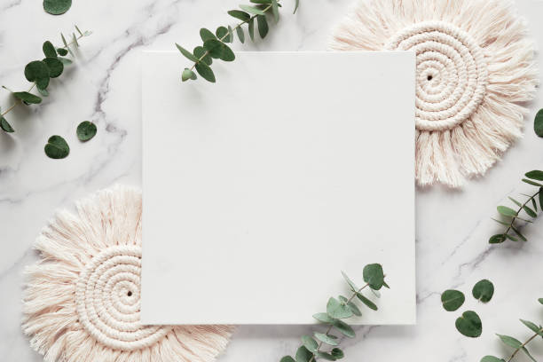 Mockup with blank square canvas, copy-space. Eucalyptus twigs, macrame pads.Top view on white marble
