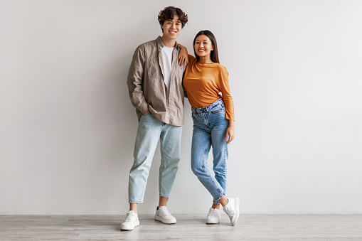 Full length portrait of happy young Asian couple in casual clothes hugging and looking at camera against white studio wall. Cheerful millennial spouses posing and smiling together