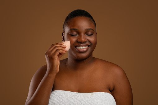 Portrait Of African American Plus Size Woman With Acne Putting On Cosmetic Foundation Base Using Sponge, Beautiful Female Applying Make Up Covering Pimples, Isolated Brown Studio Background