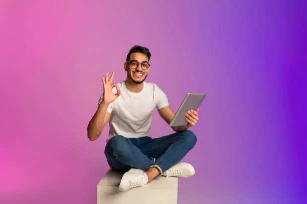Full length of young Arab guy using tablet pc, showing okay gesture, recommending new app in neon light. Millennial middle Eastern man with touch pad browsing web, working remotely