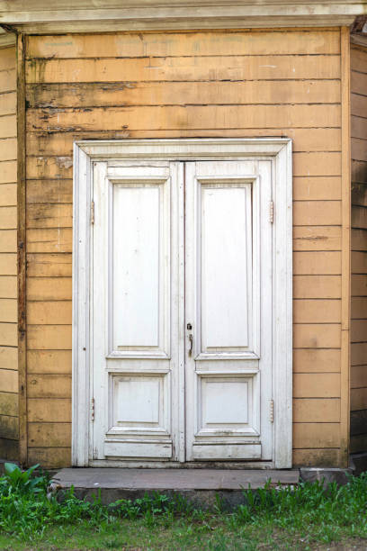 uma velha porta branca de folhas duplas em uma velha casa de madeira. a fachada está em baia com tábuas amarelas. placas são fortemente intemperado e envelhecido - weatherd - fotografias e filmes do acervo