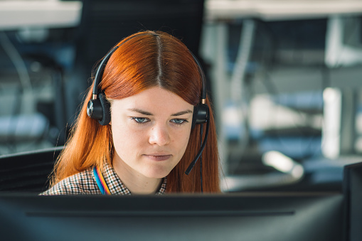 Beautiful young Caucasian red hair girl dressing casual working in the office or class at the desk in front of two monitors. Focus on job, concentration and study concept